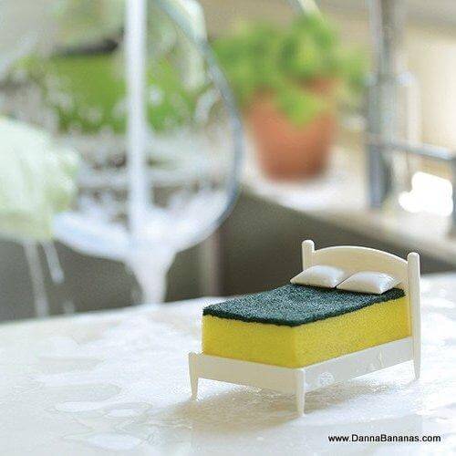 The Clean Dreams Kitchen Sponge Holder by Danna Bananas showcases a creative design where a yellow cleaning sponge serves as the mattress and a green scrubber acts as the bedsheet. The set is completed with two small white pillows, all creatively imagined against the backdrop of a softly blurred plant, adding an extra touch of charm.