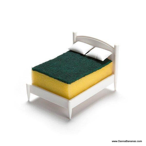 The Clean Dreams Kitchen Sponge Holder by Danna Bananas resembles a miniature bed with two white pillows and a sponge mattress, featuring a yellow base and dark green top. This charming display piece adds quirky functionality to your space as it rests on a plain white background, while also being dishwasher safe.