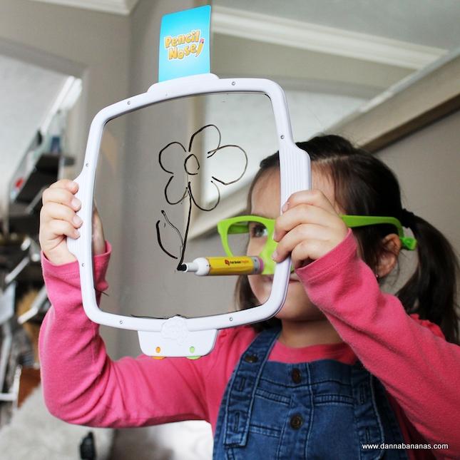 A child with green glasses and a pink long-sleeve shirt draws a flower on a transparent board, capturing the playful spirit of family games such as Danna Bananas' Pencil Nose Game. The background showcases a cozy room with stairs and furniture, suggesting lively gatherings and joyful moments.