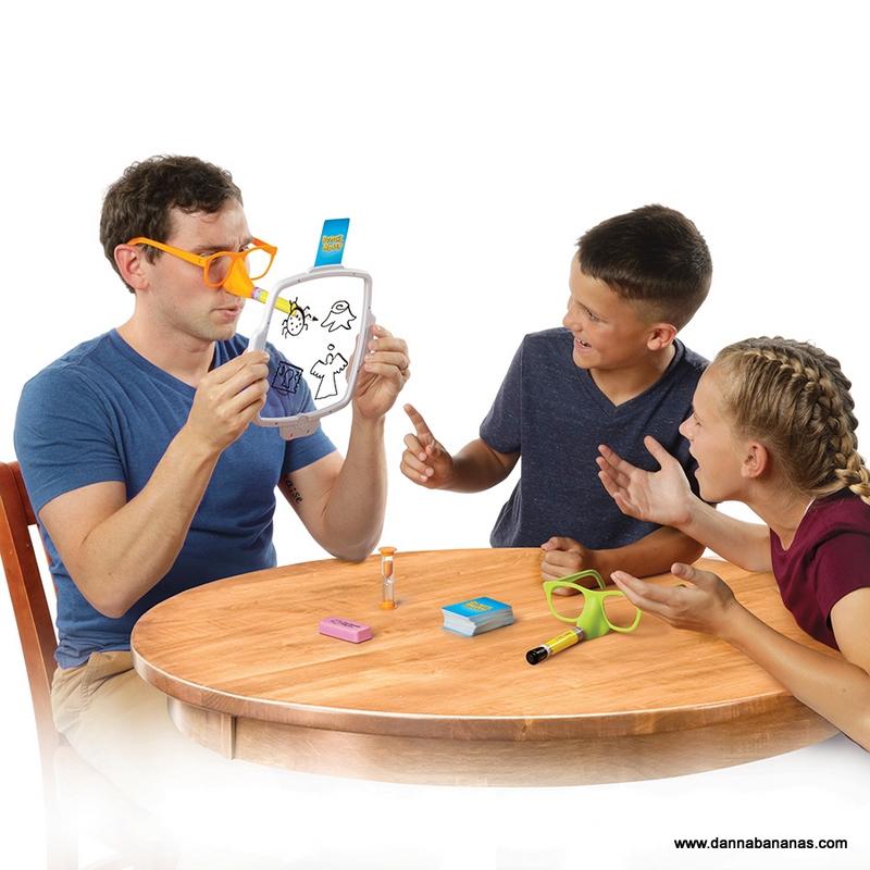 Three people are gathered around a table, deeply engaged in the Pencil Nose Game by Danna Bananas. One person, wearing glasses, holds a transparent board filled with sketches as the other two observe and point excitedly. The table is scattered with various party game items like cards and a sand timer.