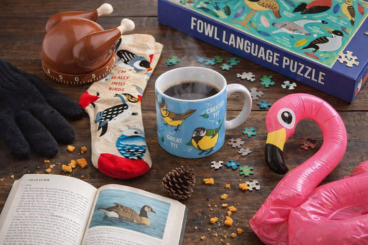 Funny bird lover gift flat lay featuring Calm Your Tits mug with Great Tit and Crested Tit birds, I'm Really Into Birds socks, Fowl Language puzzle, Gobble Getter Turkey Timer, and giant flamingo float.