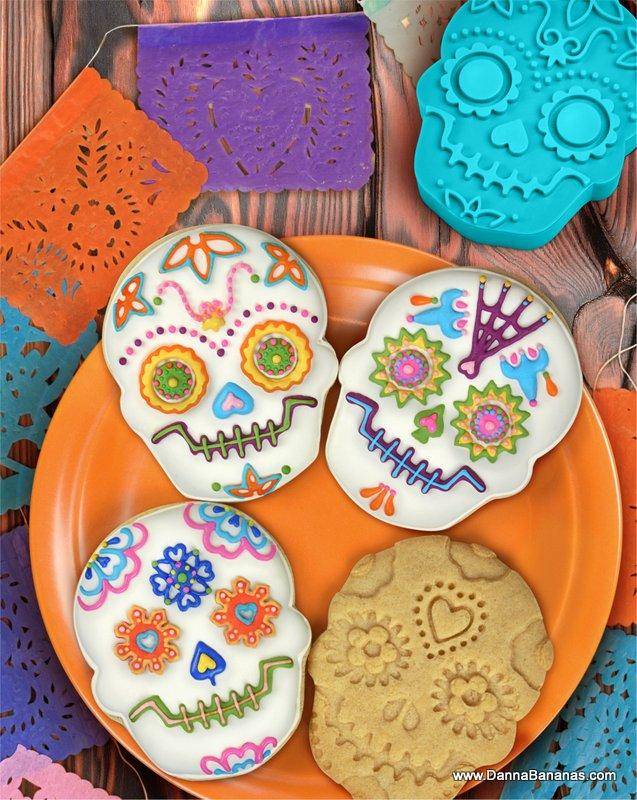 Colourful sugar skull cookies decorated with vibrant icing on an orange plate, surrounded by papel picado banners — a festive Day of the Dead treat from Danna Bananas