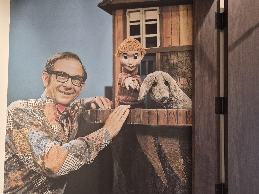 Ernie Coombs smiling in glasses and a patterned shirt, leaning on the Mr. Dressup set beside the Casey and Finnegan puppets.