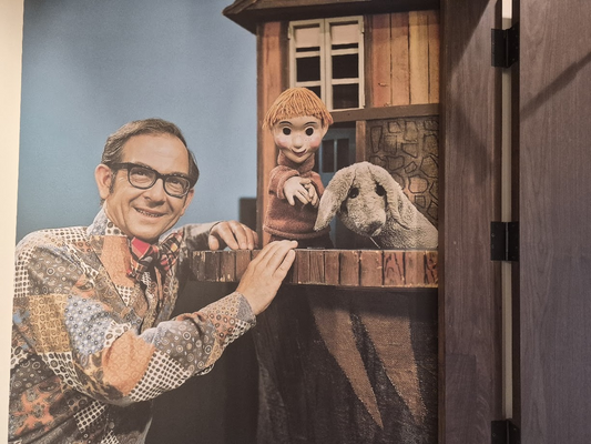 Ernie Coombs smiling in glasses and a patterned shirt, leaning on the Mr. Dressup set beside the Casey and Finnegan puppets.
