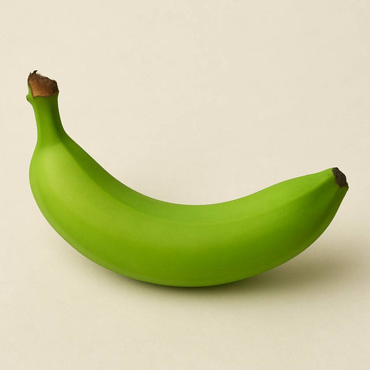 Close-up of a single green unripe banana on a neutral background, symbolizing growth, patience, and the almost-ready stage of Danna Bananas’ journey.