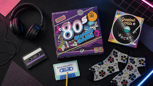 Retro gifts flat lay with an Awesome 80s Board Game, cassette sticky notes, vinyl record socks and wired headphones.