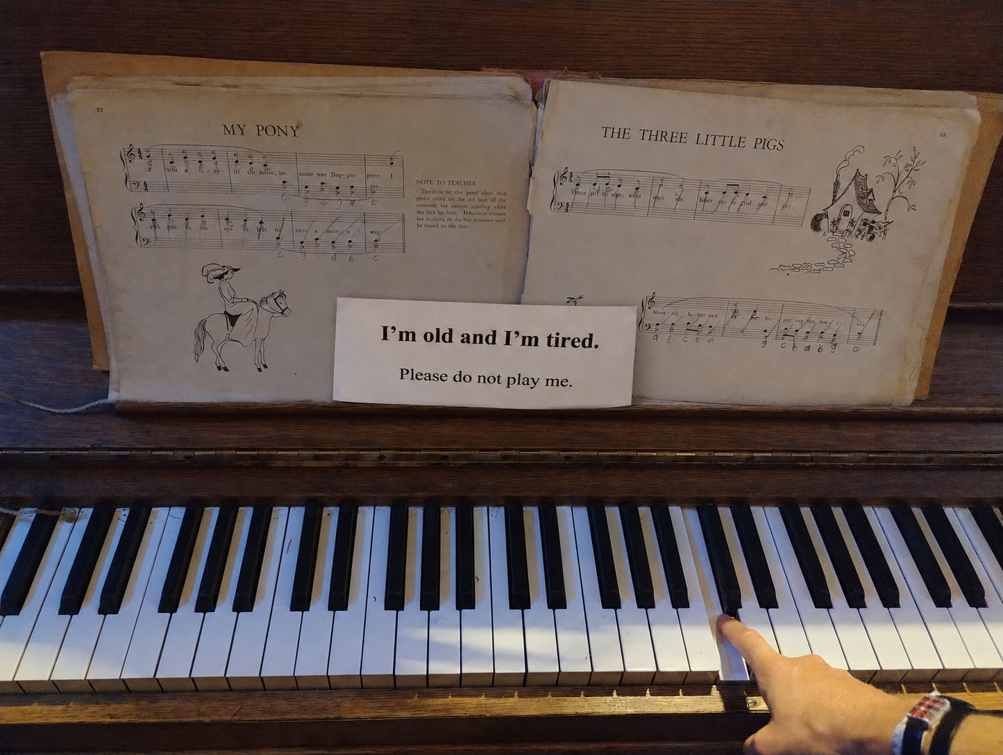 Old upright piano with an open vintage songbook and a sign that reads, I’m old and I’m tired. Please do not play me..