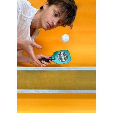 Person playing Desktop Pickleball with miniature paddle and ball over office desk net against a bright orange background.