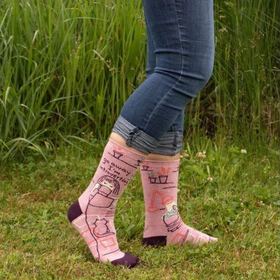 Go Away I’m Introverting funny novelty socks by Danna Bananas, shown being worn outdoors with rolled jeans, featuring a cozy pink and purple design with a cat and tea theme.