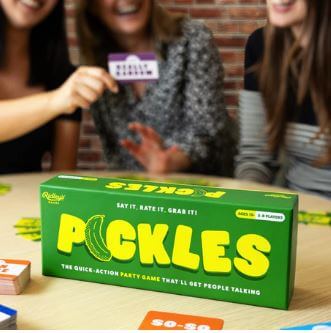 Three people are seated around a table, engrossed in a game of “Pickles The Quick Action Party Game” by Danna Bananas. The game box is prominently displayed in the foreground, featuring bright green and yellow colors with a playful pickle graphic. One person holds a game card, while everyone is smiling and fully engaged.