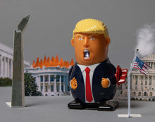 Trumpzilla wind-up toy sparking from its mouth, with a red winding key, posed before a miniature burning White House, cracked Washington Monument and American flag.