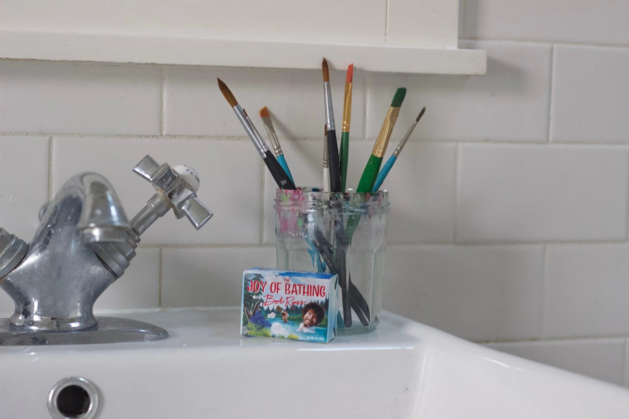 The Joy of Bathing Bob Ross Soap on the back of the sink with a jar of paint brushes