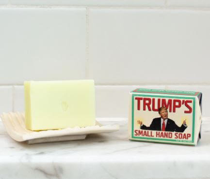 A bar of Danna Bananas' "Trump's Small Hand Soap" rests on a dish next to its novelty box, featuring a cartoon figure. This cucumber olive oil scented gift adds humor and refreshment to the scene against white tiles.