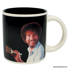 The Bob Ross Self Painting Mug by Danna Bananas features a black mug with an image of a smiling person with curly hair and a paintbrush, reminiscent of Bob Ross. The plain background accentuates the design, making it an ideal self-painting or disappearing mug for art lovers.