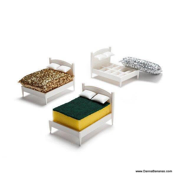 Three miniature beds with white frames are displayed. One bed features a glittery gold cover, another has a dark green cover with a yellow base, and the third holds the Danna Bananas Clean Dreams Kitchen Sponge Holder, which includes a silver kitchen sponge resembling a mattress, making it both functional and stylish as it rests in its charming little holder.