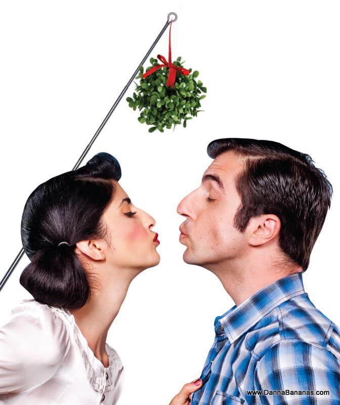 A woman and a man are about to kiss while standing under a Danna Bananas Extend-Mistletoe hanging from a rod. The woman, with dark hair styled in an updo and wearing a white blouse, faces the man, who has short dark hair and is dressed in a blue plaid shirt. The image captures the playful and festive tone of the Christmas season.