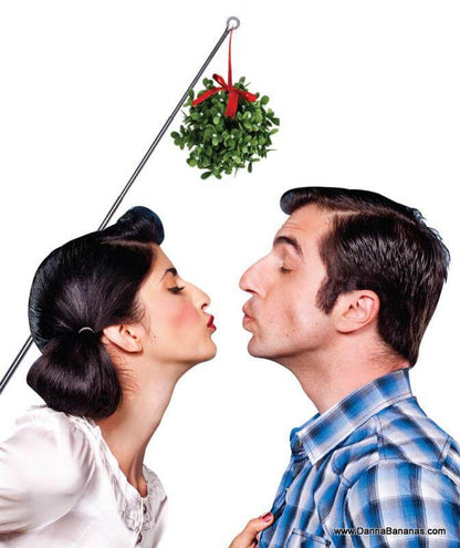 A woman and a man are about to kiss while standing under a Danna Bananas Extend-Mistletoe hanging from a rod. The woman, with dark hair styled in an updo and wearing a white blouse, faces the man, who has short dark hair and is dressed in a blue plaid shirt. The image captures the playful and festive tone of the Christmas season.
