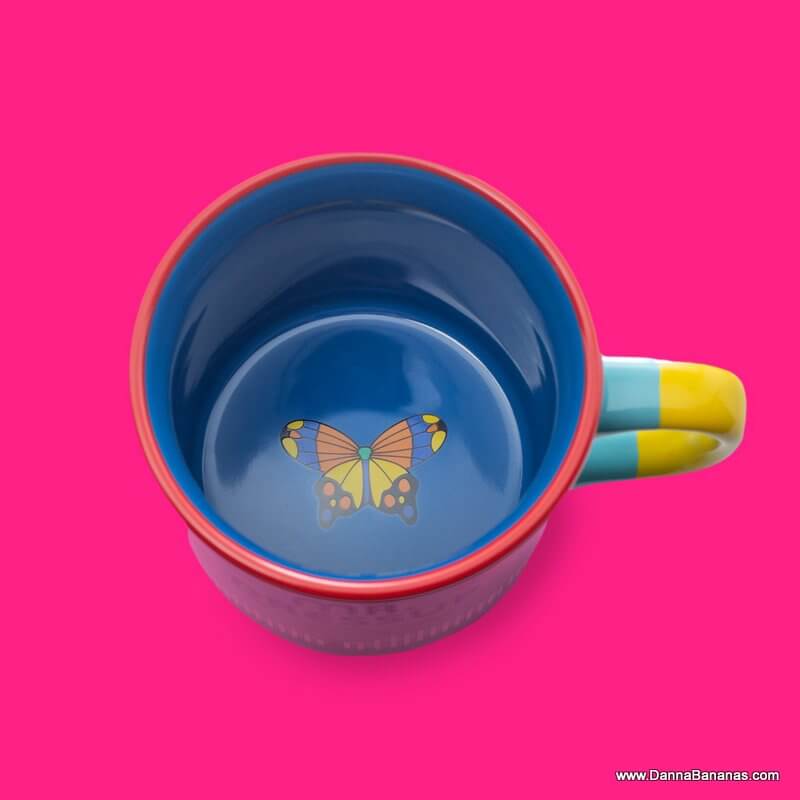 Mr. Dressup ceramic mug seen from above, showing a colourful butterfly printed on the blue interior base, red rim and yellow-accented handle.