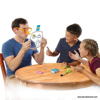 Three people are gathered around a table, deeply engaged in the Pencil Nose Game by Danna Bananas. One person, wearing glasses, holds a transparent board filled with sketches as the other two observe and point excitedly. The table is scattered with various party game items like cards and a sand timer.
